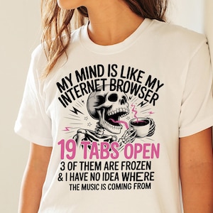 Puede incluir: Camiseta blanca con un diseño gráfico que presenta un esqueleto sosteniendo una taza de café, con el texto "MY MIND IS LIKE MY INTERNET BROWSER" y "19 TABS OPEN". Texto adicional: "3 OF THEM ARE FROZEN & I HAVE NO IDEA WHERE THE MUSIC IS COMING FROM".