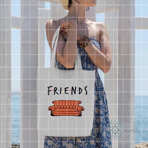 May include: White tote bag featuring the word "FRIENDS" in black and the iconic orange couch from the TV show. The bag is held by a person wearing a blue dress, with a beach and white structure in the background.