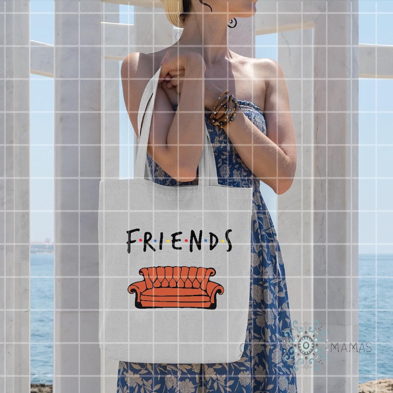 May include: White tote bag featuring the word "FRIENDS" in black and the iconic orange couch from the TV show. The bag is held by a person wearing a blue dress, with a beach and white structure in the background.