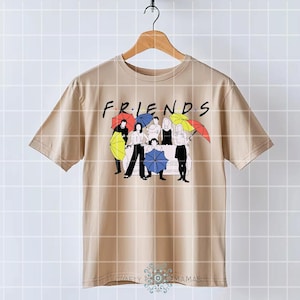May include: Tan t-shirt featuring the word "FRIENDS" in a black font. Below the word, there is a graphic of six people with umbrellas in various colors. The shirt is hanging on a wooden hanger.