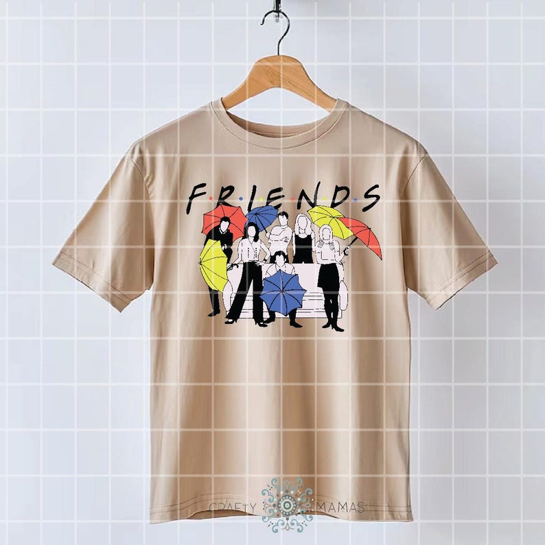 May include: Tan t-shirt featuring the word "FRIENDS" in a black font. Below the word, there is a graphic of six people with umbrellas in various colors. The shirt is hanging on a wooden hanger.