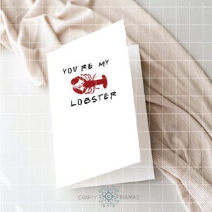 May include: A white greeting card with the text "YOU'RE MY LOBSTER" in black, with a red lobster illustration. The card is folded and resting on a white surface, with a beige fabric in the background.
