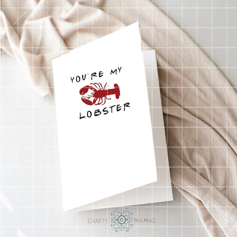 May include: A white greeting card with the text "YOU'RE MY LOBSTER" in black, with a red lobster illustration. The card is folded and resting on a white surface, with a beige fabric in the background.