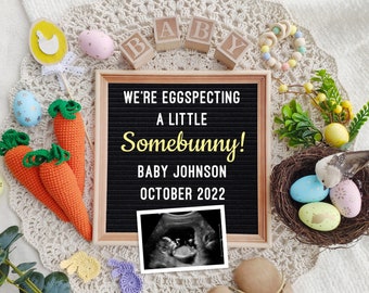 May include: A black letter board with the text "We're eggspecting a little Somebunny! Baby Johnson October 2022". The board is surrounded by Easter decorations, including carrots, eggs, and a bird's nest.