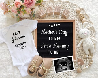 May include: A white onesie with the text "BABY Johnson DECEMBER -2022-" on it, a letter board with the text "HAPPY Mother's Day TO ME! I'm a Mommy TO BE!" and a framed ultrasound image. The onesie is surrounded by a white crocheted blanket, a white bunny, wooden blocks, and a wooden teether.