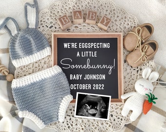 May include: A baby announcement with a chalkboard sign that reads "We're eggspecting a little somebunny!" surrounded by baby items. Includes a crochet bunny hat, shorts, booties, a rattle, a stuffed bunny, and an ultrasound image.