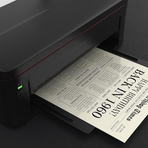May include: A black printer with a white sheet of paper emerging from it. The paper has a newspaper design with the text "The Birthday Times" and "Happy Birthday! Back in 1960".