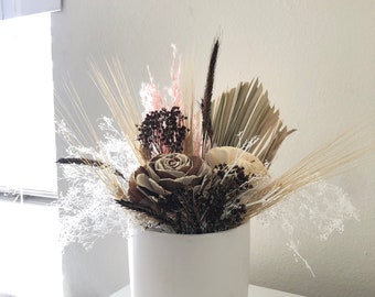May include: A white ceramic vase filled with dried flowers, including a large, light brown wooden rose, smaller white wooden roses, and dried grasses in shades of brown and beige.