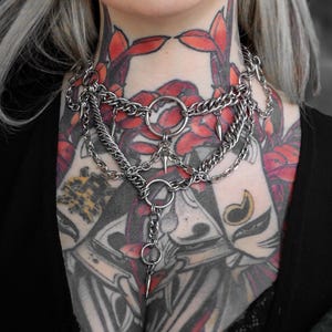 May include: A silver-toned layered chain necklace with multiple strands and a central ring design. The necklace features dangling spikes and various chain styles, creating a bold, edgy aesthetic. The jewelry is worn on a person with visible neck tattoos.