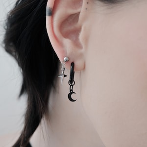 May include: A person wearing a black choker necklace with a small silver pendant. They have a black hoop earring with a dangling crescent moon charm and a silver star charm.