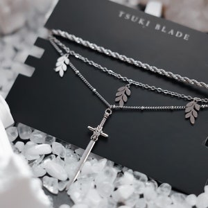 May include: A silver necklace featuring a sword pendant and leaf charms. The necklace is presented on a black card with the text "TSUKI BLADE".