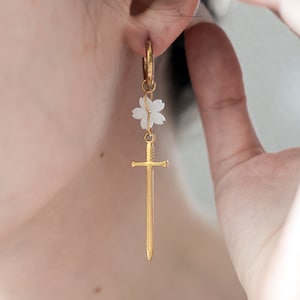 May include: Gold hoop earrings with a white flower charm and a gold sword dangle.
