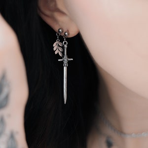 May include: Silver earrings with a leaf and sword design. The earrings hang from the ear. The image is a close-up of the earrings and the person's ear. The person is wearing other silver jewellery.