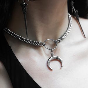 May include: A silver-toned choker necklace with a crescent moon pendant. The necklace features a chain design and a large circular clasp. Matching dagger-shaped earrings are also visible.