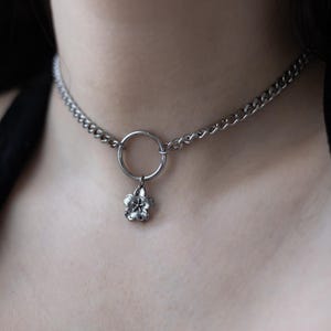 Op de afbeelding: Zilverkleurige choker ketting met een ketting en een grote cirkelvormige hanger. Een kleine bloemhanger hangt aan de cirkel. De ketting wordt om de nek gedragen.
