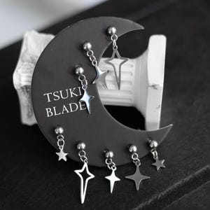 以下が含まれることがあります： 6つの銀色の星型のイヤリングのセットで、チェーンがぶら下がっています。イヤリングは、黒い三日月型のカードに表示されており、カードには「TSUKI BLADE」という文字が印刷されています。