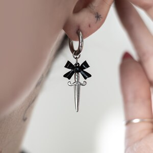 May include: Silver hoop earring with a black bow and a silver dagger charm hanging from it.