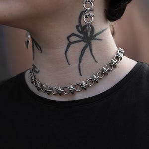 May include: Silver-coloured choker necklace with conical spikes and matching earrings. The earrings are made of linked rings and a conical pendant. The jewellery is worn with a black top. The image features a tattoo of a spider on the neck.