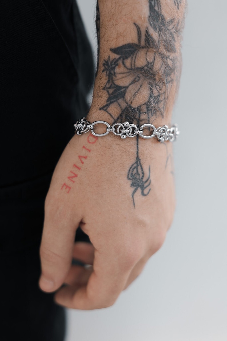 May include: A silver chain bracelet with a decorative design. The bracelet is on a person's wrist with a tattoo of a spider and a flower.