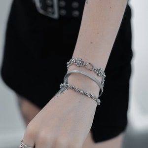 May include: A person's wrist wearing a set of four silver bracelets, including a chain link bracelet, a rope chain bracelet, and two thin bangle bracelets. The person is also wearing several silver rings on their fingers.