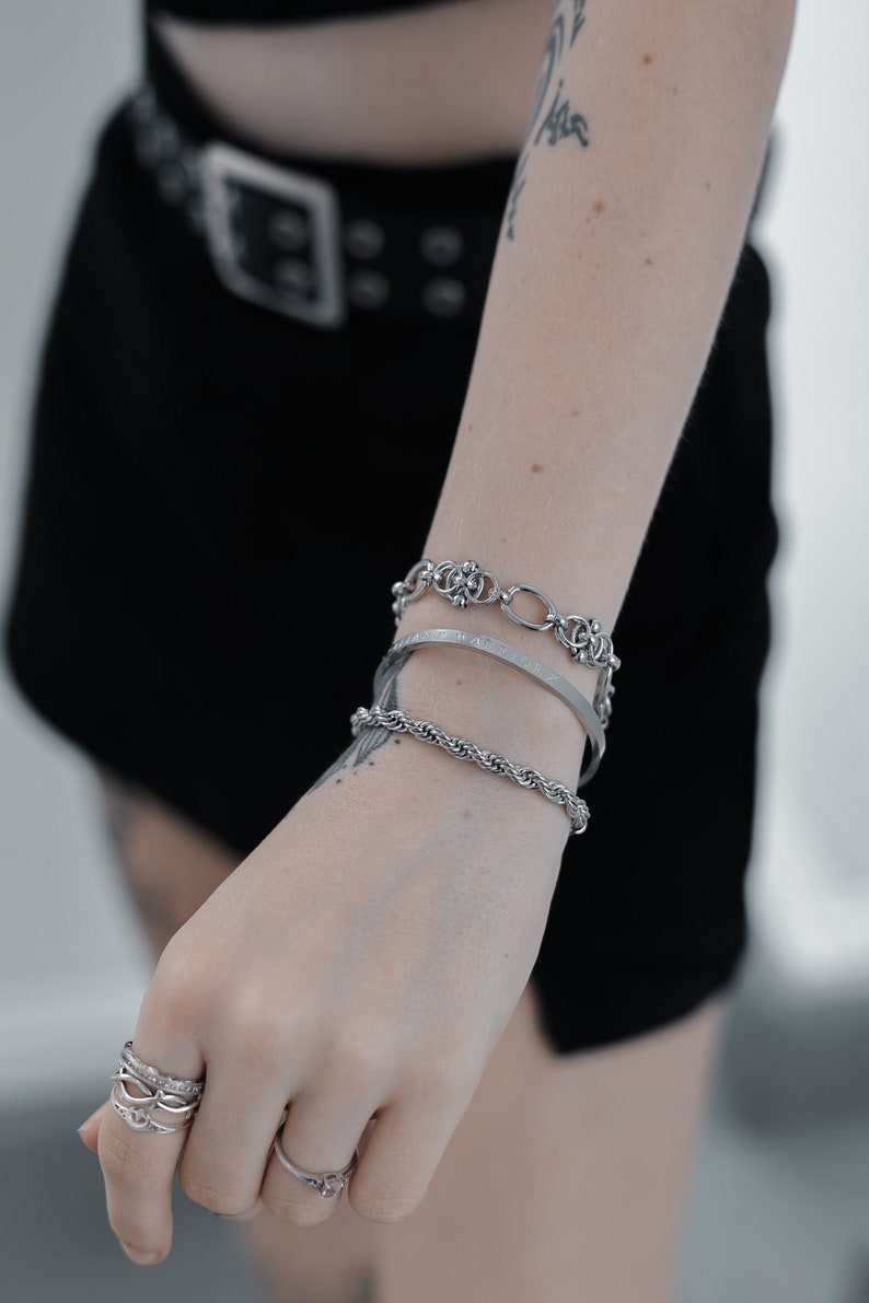 May include: A person's wrist wearing a set of four silver bracelets, including a chain link bracelet, a rope chain bracelet, and two thin bangle bracelets. The person is also wearing several silver rings on their fingers.