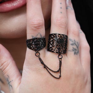 May include: Black metal ring set connected by a chain. The rings feature intricate openwork designs. One ring is on the index finger, the other on the middle finger. The chain hangs between the rings.