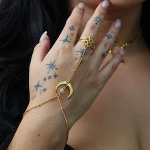 May include: A gold hand chain featuring a crescent moon pendant and a fine chain linking to a ring. The hand chain is worn on a hand adorned with star tattoos and a matching ring with an elaborate design.