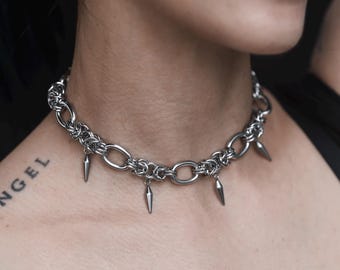 Puede incluir: Un collar tipo gargantilla de metal plateado que presenta una serie de eslabones circulares entrelazados y segmentos de malla de cadena. El collar está adornado con cuatro pequeños dijes lisos en forma de punta que cuelgan.