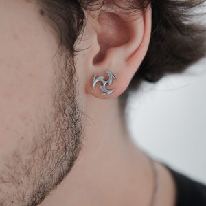 Könnte beinhalten: Nahaufnahme eines silberfarbenen Ohrsteckers mit einem dreizackigen Design. Der Ohrring hat die Form einer stilisierten Spirale oder eines Wirbels. Der Ohrring wird am Ohr einer Person mit Bart und dunklem Haar getragen.