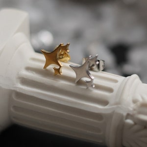 Pode incluir: Um par de brincos em forma de estrela, um dourado e outro prateado, sobre uma coluna branca.