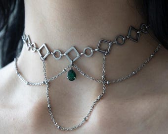 May include: A silver-toned choker necklace featuring a series of connected diamond-shaped and circular metal links. A delicate chain drapes below the main band, accented by a single, small, teardrop-shaped dark green gemstone pendant at the centre.