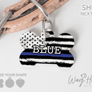 May include: A white bone-shaped dog tag with a blue line and an American flag design. The tag says "BLUE" in black letters. The tag is attached to a brown leather dog collar.