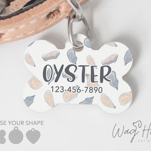 May include: White bone-shaped dog tag with a blue and beige oyster pattern. The tag says "Oyster" and "123-456-7890".