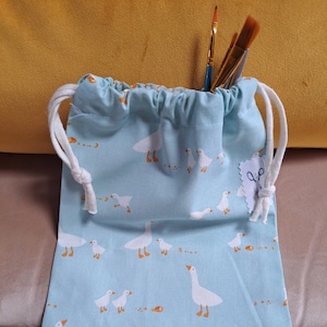 Può includere: Una borsa a cordoncino blu chiaro con un motivo di oche bianche. La borsa ha un cordoncino bianco e una piccola etichetta bianca con un paio di forbici.