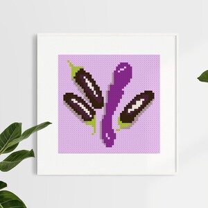 May include: A framed cross-stitch artwork featuring a pixelated design of eggplants. The eggplants are in shades of purple, brown, and green, set against a light purple background. The artwork is displayed in a white frame.