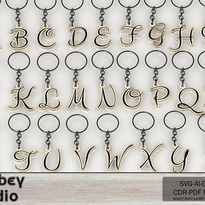 May include: A collection of gold-colored alphabet keychains, each letter suspended from a black chain and keyring. The letters are in a cursive font. The image also includes the text "Atabey studio" and "SVG-AI-DXF CDR-PDF FILES READY EASY LASER CUT FILES".