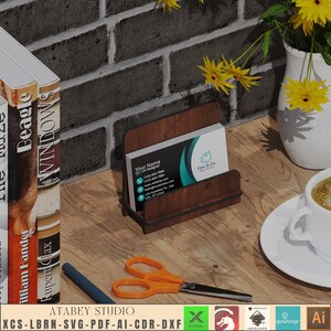 May include: A wooden business card holder with a brown wood grain finish. The holder is sitting on a wooden table with a stack of books and a vase of yellow flowers in the background. The business card holder has a business card inside with the text "Your Name UI/UX Designer Your & Co. Real Estate" printed on it.