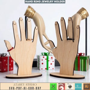 Op de afbeelding: Houten handvormige ringhouders met ringen aan de vingers. De houders zijn lichtbruin met een donkerbruine basis. De achtergrond bevat een decoratieve hand en geschenkdozen. De tekst "HAND RING JEWELRY HOLDER" en "ATABEY STUDIO SVG-PDF-AI-CDR-DXF" zijn zichtbaar.