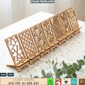 May include: A set of seven light-colored wooden phone stands featuring various laser-cut geometric and organic patterns. Each stand has a small horizontal shelf and a base. Text on the image includes STAND TEMPLATES, 3mm, 1/8in, and ATABEY STUDIO.