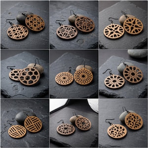 May include: A collection of wooden earrings with various geometric designs. The earrings are light brown with black ear wires. The designs include circles, hexagons, and other intricate patterns. The earrings are displayed on a dark gray surface.
