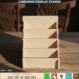 Puede incluir: Un expositor de madera de cuatro niveles, ideal para exhibir art&iacute;culos. El expositor est&aacute; hecho de madera de color claro y presenta el texto "2-DESIGNS DISPLAY STANDS" en la parte superior. El expositor tiene 6 mm de grosor. El logotipo de Atabey Studio es visible.