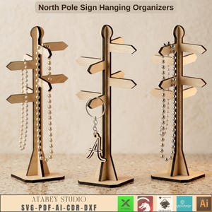 May include: Three light-coloured wooden signpost-style jewellery organisers with multiple horizontal arms. The text North Pole Sign Hanging Organisers appears at the top. Two organisers hold beaded necklaces, and one holds a metal letter A keychain.