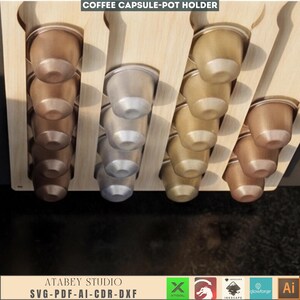 Puede incluir: Soporte de madera para c&aacute;psulas de caf&eacute; con m&uacute;ltiples ranuras, lleno de varias c&aacute;psulas de caf&eacute; met&aacute;licas. El soporte est&aacute; etiquetado como "COFFEE CAPSULE-POT HOLDER" y "ATABEY STUDIO SVG-PDF-AI-CDR-DXF".