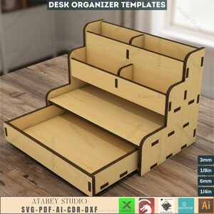 May include: A wooden desk organizer with multiple compartments and a pull-out drawer. The organizer has a tiered design with various sections for storing office supplies. The text "DESK ORGANIZER TEMPLATES" is at the top. The organizer is made of light-colored wood with dark brown edges.