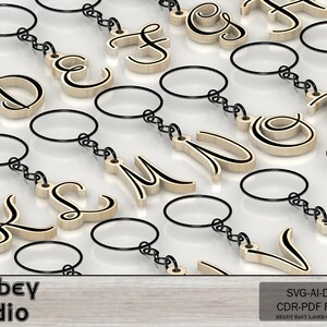May include: Wooden keychains with various capital letters in a cursive font, each with a black outline. The keychains have a silver-colored chain and a round key ring. The background is a light, reflective surface. The image also includes the text "Atabey studio".