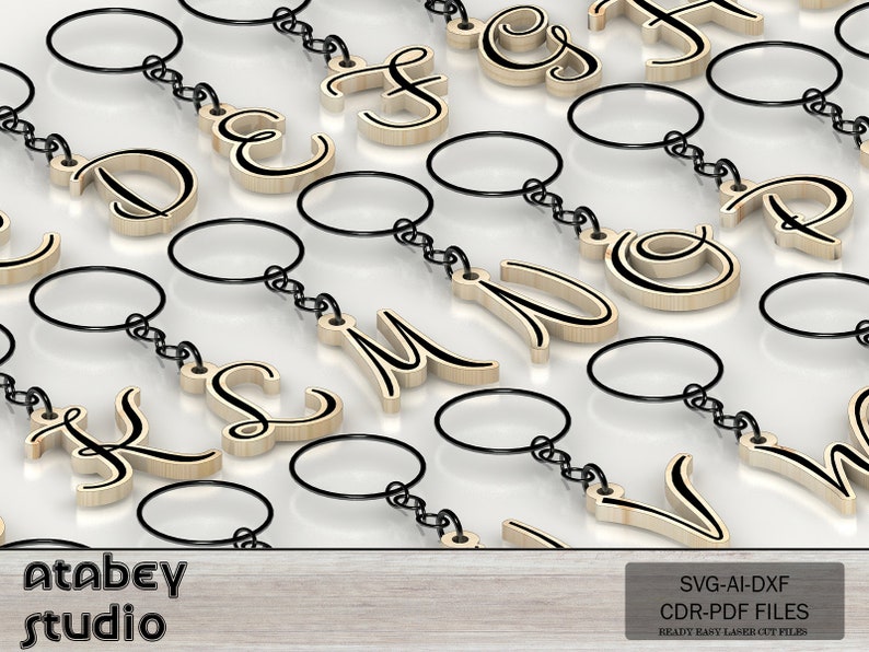 May include: Wooden keychains with various capital letters in a cursive font, each with a black outline. The keychains have a silver-colored chain and a round key ring. The background is a light, reflective surface. The image also includes the text "Atabey studio".