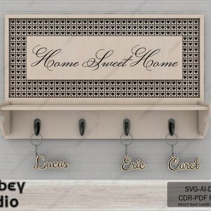 Può includere: Un porta chiavi in legno con uno scaffale e tre ganci. Il pannello superiore presenta un design a forma di cuore e il testo "Home Sweet Home". Tre portachiavi in legno con i nomi "Lucas", "Eric" e "Carol" sono appesi ai ganci.