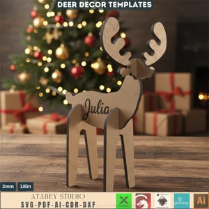 Puede incluir: Decoración de ciervo de madera con el nombre "Julia" inscrito en un lado. El ciervo es de madera marrón claro con bordes negros y está frente a un árbol de Navidad. El texto "DEER DECOR TEMPLATES" está en la parte superior.
