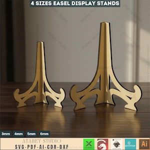 Op de afbeelding: Twee goudkleurige ezel display stands op een houten oppervlak. De stands hebben een decoratieve basis en een hoge, puntige top. De tekst "4 SIZES EASEL DISPLAY STANDS" staat bovenaan. De tekst "ATABEY STUDIO" en "SVG-PDF-AI-CDR-DXF" staan onderaan.