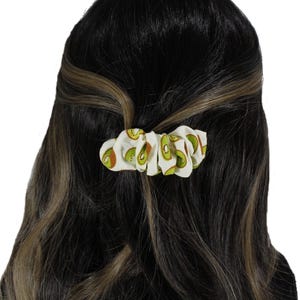Puede incluir: Una goma para el pelo blanca con un estampado repetido de rodajas de kiwi verdes y marrones. La goma para el pelo se lleva en el pelo, con reflejos castaños oscuros y rubios visibles.
